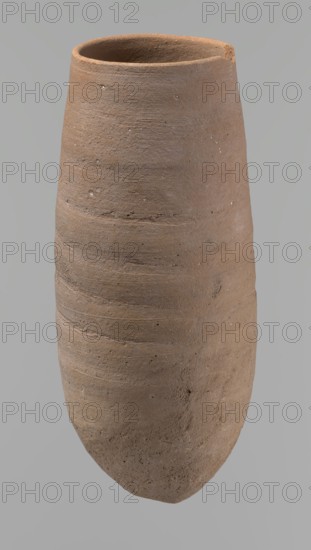 Vessel, Vessel with a conical shape and a pointed bottom. Straight, tapering rim. Made on the wheel of Nile clay type NB2 Vienna System classification. Bottom and outer surface worked with a hard tool. Surface even. Light brown. Fairly well preserved, with minor damage on the rim. c. 1550–c. 1069 BC, New Kingdom, Deir el-Medina. Vessel; height 20.8 cm; diameter 7 cm lip, 9 cm body., A tall, narrow ceramic vessel with a rounded base and an open rim, shown against a plain gray background; the vessel has a textured, slightly rough surface with horizontal ridges and small pits, and its color ranges in shades of brown and tan with subtle darker and lighter variations.