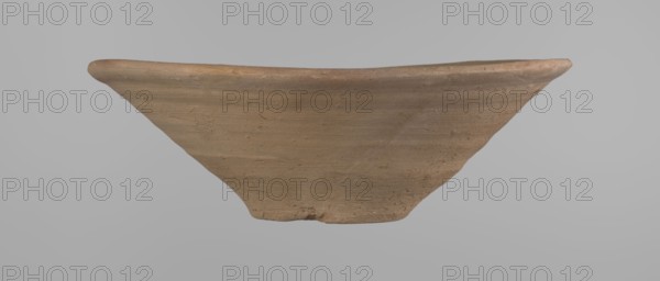 Bowl, Bowl with a conical shape and a flat bottom, slightly asymmetrical. Straight, flaring rim. Vessel made on the wheel of Nile clay type NB2 according to the Vienna System classification. The bottom is cut off with a knife. Surface leveled. The outer part of the vessel is finished to approximately half its height with a knife or similar tool. Color of the vessel: yellow-red. Vessel reconstructed from two original fragments. c. 1550–c. 1069 BC; New Kingdom Ramesside period?, Deir el-Medina. Vessel. Height 9.2 cm; diameter 27.8 cm rim; diameter 8.4 cm bottom., A fragment of a shallow, wide-rimmed ceramic dish in shades of brown and tan is shown against a neutral gray background; the piece has a curved rim with a slightly raised edge and a textured surface with visible horizontal striations and small imperfections, and the fragment tapers toward a narrower base with irregular, worn edges.