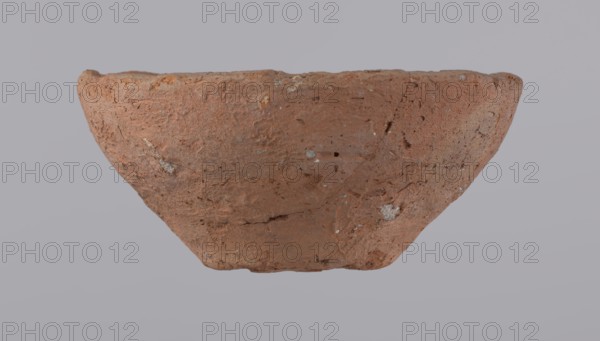 Bowl, probably votive, with a conical shape and a flat bottom. Straight rim, slightly curled towards the center. Wheel-made vessel of Nile clay type NC according to the Vienna System classification. Leveled surface. Flat, irregular bottom, cut off with a string. Vessel color: brown-red. Vessel reconstructed from three original fragments. Rim slightly damaged. c. 1550–c. 1069 BC; New Kingdom, Deir el-Medina. Vessel, height 4.8 cm, diameter 10.2 cm lip, diameter 4 cm bottom., A roughly bowl-shaped object with a textured, worn surface occupies the center against a plain background; the object is predominantly reddish-brown with small areas of lighter grayish-white flecks and patches, showing rough, uneven edges and visible pits and scratches across its exterior, and the background appears as a muted pale gray.