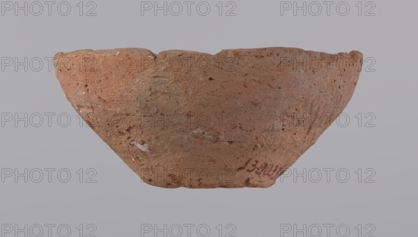 Bowl, probably votive, with a conical shape and a flat bottom. Straight rim, slightly curled towards the center. Wheel-made vessel of Nile clay type NC according to the Vienna System classification. Leveled surface. Flat, irregular bottom, cut off with a string. Vessel color: brown-red. Vessel reconstructed from three original fragments. Rim slightly damaged. c. 1550–c. 1069 BC; New Kingdom, Deir el-Medina. Vessel, height 4.8 cm, diameter 10.2 cm lip, diameter 4 cm bottom, A roughly textured, shallow bowl-shaped object sits against a pale gray background; the object has an uneven rim and a coarse surface with visible pits and small inclusions, showing a dominant reddish-brown clay color with patches of lighter beige and small flecks of gray and white, and a few areas of darker brown; the shape tapers toward the base and the rim edge is irregular and worn.