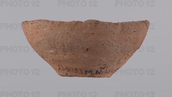 Bowl, probably votive, conical shape with a flat bottom. Straight rim, slightly curled toward the center. Wheel-made vessel of Nile clay type NC Vienna System. Leveled surface. Flat, irregular bottom, cut off with a string. Vessel color: brown-red. Reconstructed from three original fragments; rim slightly damaged. c. 1550–c. 1069 BC, New Kingdom, Deir el-Medina. Vessel; height 4.8 cm, diameter 10.2 cm lip, diameter 4 cm bottom., A roughly semicircular, shallow clay bowl with a textured, pitted surface and an uneven rim sits against a pale gray background; the bowl is predominantly reddish-brown with areas of darker brown and small flecks of lighter tan, showing visible roughness and tiny inclusions in the clay, and the inner and outer surfaces display variations in tone and subtle scratches and chips along the rim.