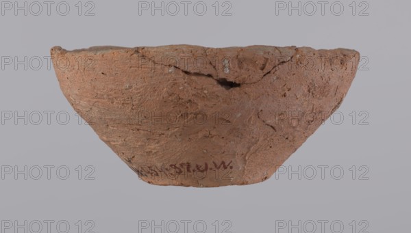 Bowl, probably votive, with a conical shape and a flat bottom. Straight rim, slightly curled towards the center. Wheel-made vessel of Nile clay type NC Vienna System. Leveled surface. Flat, irregular bottom, cut off with a string. Vessel color: brown-red. Reconstructed from three original fragments; rim slightly damaged. c. 1550–c. 1069 BC, New Kingdom; provenance: Deir el-Medina. Vessel; height 4.8 cm; diameter 10.2 cm lip; diameter 4 cm bottom., A roughly bowl-shaped broken clay object with an uneven rim and a textured, cracked surface in reddish-brown tones, showing areas of lighter and darker brown and small pale flecks; it sits against a smooth pale gray background.