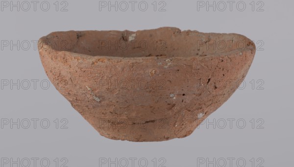 Bowl, probably votive, conical shape with flat bottom. Straight rim, slightly curled toward center. Wheel-made vessel of Nile clay type NC Vienna System. Leveled surface. Flat, irregular bottom, cut off with a string. Brown-red color. Reconstructed from three original fragments; rim slightly damaged. c. 1550–c. 1069 BC, New Kingdom, Deir el-Medina. Vessel; height 4.8 cm, diameter 10.2 cm lip, diameter 4 cm bottom., A roughly shaped, shallow bowl with an uneven rim sits centered against a pale gray background; the bowl has a coarse, textured surface showing a mix of earthy brown and reddish-brown tones with small patches of lighter beige and gray, tiny pits and rough inclusions, and irregularities along its interior and exterior surfaces.