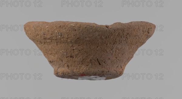 Bowl, probably votive, conical shape with flat bottom. Irregular walls; straight, slightly flaring rim; irregular bottom, probably cut off with a string. Wheel-made vessel of Nile clay type NC Vienna System. Surface only slightly leveled. Vessel color: brown. Well-preserved with minor surface erosion. c. 1550–c. 1069 BC New Kingdom, Deir el-Medina. Vessel; height 3.7 cm; diameter 8.2 cm lip; diameter 4.2 cm bottom., A rough textured, shallow bowl-shaped object with an irregular rim, shown against a gray background; the surface is coarse and porous with small pits and inclusions, and the visible colors are various shades of brown with a few darker brown flecks and a small area of muted red near the base.
