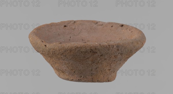 Bowl, probably votive, conical shape with a flat bottom. Irregular walls; straight, slightly flaring rim; irregular bottom probably cut off with a string. Wheel-made vessel of Nile clay type NC Vienna System. Surface only slightly leveled. Vessel color brown. Well-preserved with minor surface erosion. c. 1550–c. 1069 BC New Kingdom, Deir el-Medina. Ceramic vessel bowl. Height 3.7 cm; diameter 8.2 cm lip, 4.2 cm bottom., A single shallow bowl-shaped object made of rough, coarse material is centered against a plain light gray background; the object has an irregular rim and uneven surface texture with small pits and inclusions, showing overall earthy brown and reddish-brown tones with subtle darker speckles and patches, and the interior appears slightly smoother but still textured in similar brown and muted red hues.