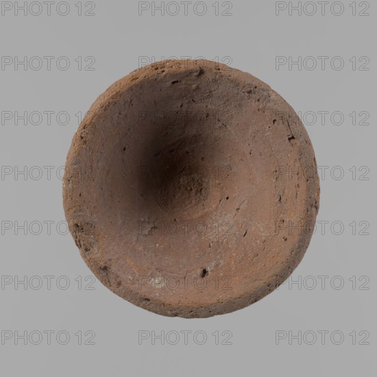 Bowl, probably votive, conical shape with a flat bottom. Irregular walls; straight, slightly flaring rim; irregular bottom, probably cut off with a string. Wheel-made vessel of Nile clay type NC Vienna System classification. Surface only slightly leveled. Vessel color brown. Well-preserved with only minor surface erosion. c. 1550–c. 1069 BC New Kingdom, from Deir el-Medina. Vessel; height 3.7 cm, diameter 8.2 cm lip, diameter 4.2 cm bottom., A roughly circular, shallow bowl-like object viewed from above with a textured, uneven surface showing small pits and chips, predominantly brown with variations of reddish-brown and some darker brown areas, set against a plain light gray background.