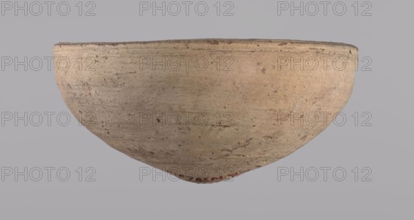 Bowl. Bowl with a conical shape and a pointed bottom. Flat, straight rim. Vessel made on the wheel of Nile clay type NB2 according to the Vienna System classification. Surface even, irregularly covered with engobe inside and outside. Color of vessel: cream. Vessel fairly well preserved, slightly damaged in the upper part. c. 1550–c. 1069 BC; New Kingdom, Deir el-Medina. Vessel, height 6.5 cm, diameter 13.6 cm lip., A worn shallow bowl with a rounded base and flared rim, showing a rough, textured surface with small pits and abrasions; the bowl is primarily beige with areas of tan and light brown, and there are subtle darker brown speckles and marks across its exterior; the background is a flat, muted gray.