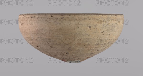 Bowl, Bowl with a conical shape and a pointed bottom. Flat, straight rim. Vessel made on the wheel of Nile clay type NB2 according to the Vienna System classification. Surface even, irregularly covered with engobe inside and outside. Color of vessel: cream. Vessel fairly well preserved, slightly damaged in the upper part. c. 1550–c. 1069 BC; New Kingdom, Deir el-Medina. Vessel; height 6.5 cm, diameter 13.6 cm lip., A large rounded vessel shown from the side and slightly upside down, with a textured surface that appears rough and worn; the vessel is mainly beige with areas of tan and light brown, and it has small darker brown speckles and patches across its surface, a slightly darker rim, and a small area of muted red and a small patch of blue-gray near the bottom; the background is a uniform pale gray.