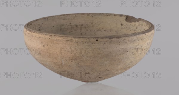 Bowl. Bowl with a conical shape and a pointed bottom. Flat, straight rim. Vessel made on the wheel of Nile clay type NB2 according to the Vienna System classification. Surface even, irregularly covered with engobe inside and outside. Color of vessel: cream. Vessel fairly well preserved, slightly damaged in the upper part. c. 1550–c. 1069 BC; New Kingdom, Deir el-Medina. Vessel, height 6.5 cm, diameter 13.6 cm lip., A shallow, rounded clay bowl with an uneven rim sits against a plain light gray background; the bowl shows a weathered, matte surface in shades of beige, tan, and light brown with darker brown speckles and small pits, a few rough spots along the rim, and subtle horizontal markings from shaping, while the interior matches the exterior colors and texture.