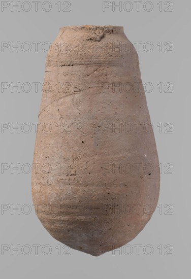 Vessel, Egyptian pottery ceramic clay vessel, New Kingdom, from Deir el-Medina; vessel height 18.5 cm, rim diameter 5.2 cm, body diameter 10.2 cm., A single large rounded vessel with a narrow opening is shown against a plain light gray background; the vessel has a rough, textured surface in shades of brown, tan, and muted reddish-brown with small darker and lighter specks and irregularities, and its form tapers slightly toward the top and bottom with subtle horizontal marks and occasional small pits and scratches visible across the exterior.
