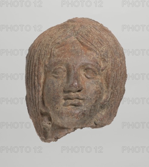 Female votive head, Female head with long, straight hair falling to the shoulders. Neck partially preserved. Elongated face with a receding forehead is characterized by wide-open eyes set shallowly under prominent eyebrow arches. The mouth is slightly open with fleshy lips, the chin protrudes forward, and full cheeks. 2nd century BC, Etruria; votive. Height 20 cm, width 12 cm, depth 8.5 cm, weight 0.94 kg., A worn sculpted head with textured hair and a neutral facial expression, rendered in earthy tones of brown and reddish-brown with patches of darker and lighter brown and some grayish areas; the hair is styled in layered, ribbed strands framing the face, the eyes are almond-shaped and recessed, the nose is rounded, and the lips are full and slightly parted; the surface shows roughness and pitting and fragments missing along the lower edge, set against a pale, light gray background.