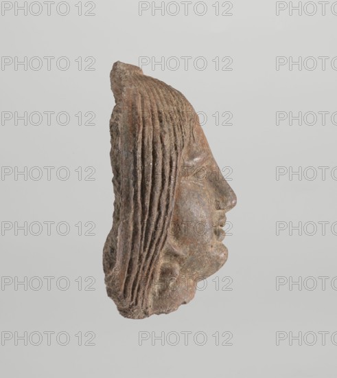 Female votive head, Female head with long, straight hair falling to the shoulders. Neck partially preserved. Elongated face with a receding forehead characterized by wide-open eyes set shallowly under prominent eyebrow arches. Mouth slightly open with fleshy lips, chin protrudes forward, and full cheeks. 2nd century BC, Etruria; votive. Height 20 cm, width 12 cm, depth 8.5 cm, weight 0.94 kg., A carved stone fragment showing the right-side profile of a face with a pronounced nose, closed lips, and a defined chin, with a section of textured hair at the back rendered as parallel grooves; the surface shows rough, weathered texture and irregular edges. The visible colors include shades of brown, reddish-brown, gray, and small areas of lighter beige. The background is a flat, pale gray.