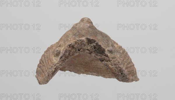 Female votive head, Female head with long, straight hair falling to the shoulders. Neck partially preserved. Elongated face with a receding forehead characterized by wide-open eyes set shallowly under prominent eyebrow arches. Mouth slightly open with fleshy lips, protruding chin, and full cheeks. 2nd century BC, Etruria; votive. Height 20 cm, width 12 cm, depth 8.5 cm, weight 0.94 kg., A weathered, arch-shaped object with a rough, textured surface in shades of brown, beige, tan, and hints of gray, showing layered ridges along its sides and an uneven, porous edge, photographed against a plain light gray background.