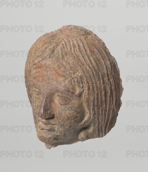 Female votive head with long, straight hair falling to the shoulders. Neck partially preserved. Elongated face with a receding forehead, wide-open eyes set shallowly under prominent eyebrow arches. Mouth slightly open with fleshy lips, protruding chin, and full cheeks. 2nd century BC, Etruria; votive. Height 20 cm, width 12 cm, depth 8.5 cm, weight 0.94 kg., A weathered sculpted head carved from a stone-like material with rough textured surfaces and visible erosion, showing a frontal face with closed or downcast eyes, a pronounced nose, full lips, and stylized wavy hair rendered in parallel grooves that frame the face and cover the top and sides of the head; the object displays an overall palette of brown, tan, reddish-brown, gray, and hints of darker brown in recessed areas, set against a plain light gray background.