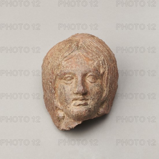 Female votive head, Female head with long, straight hair falling to the shoulders. Neck partially preserved. Elongated face with a receding forehead characterized by wide-open eyes set shallowly under prominent eyebrow arches. Mouth slightly open with fleshy lips, protruding chin, and full cheeks. 2nd century BC, Etruria; votive. Height 20 cm, width 12 cm, depth 8.5 cm, weight 0.94 kg., A weathered sculpted head with textured hair and facial features sits against a plain background; the object shows shades of brown, reddish-brown, tan, and hints of gray, with rough surface variations and small areas of lighter and darker discoloration, and a neutral light gray backdrop.