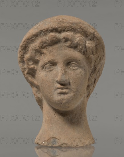 Female votive head, female head in a wreath, slightly tilted to the right shoulder. Under the wreath are visible regularly arranged locks of hair, framing a low, triangular forehead and temples. Ears are exposed. Deep-set eyes, almost flat in the elaborately worked eyelids, small nose, small mouth with fleshy lips, short, strongly defined chin, full cheeks. 3rd century BC, Etruria, votive. Height 26 cm, width 16.5 cm, depth 13.5 cm, weight 2.1 kg., A sculpted head made of brownish clay with a rounded headdress or cap and curly hair, showing a face with closed or slightly open eyes, a straight nose, and a small mouth; the surface has a few lighter and darker spots and some rough, worn texture; the neck is tapered and sits on a narrow base, and the background is a neutral gray.