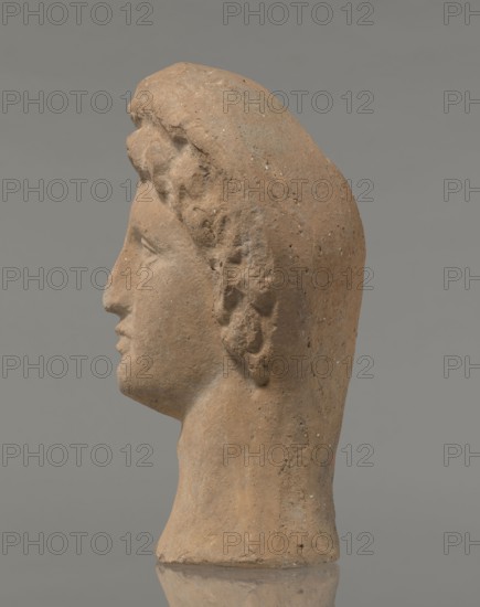 Female votive head, Female head in a wreath, slightly tilted to the right shoulder. Under the wreath are visible regularly arranged locks of hair, framing a low, triangular forehead and temples. Ears exposed. Deep-set eyes, almost flat in the elaborately worked eyelids; small nose; small mouth with fleshy lips; short, strongly defined chin; full cheeks. 3rd century BC, Etruria; votive. Height 26 cm, width 16.5 cm, depth 13.5 cm, weight 2.1 kg., A profile view of a sculpted head in muted brown and tan tones, showing a smooth forehead, defined nose, lips, and chin, with textured curly hair along the side and back of the head; the surface has small mottled speckles and rough patches, and the base of the neck is visible, all set against a flat gray background.