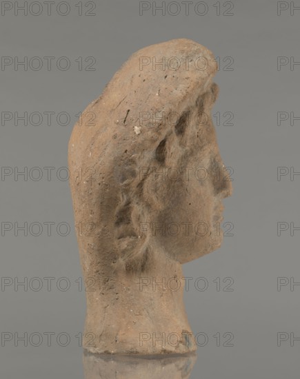 Female votive head, Female head in a wreath, slightly tilted to the right shoulder. Under the wreath are visible regularly arranged locks of hair, framing a low, triangular forehead and temples. Ears are exposed. Deep-set eyes, almost flat in the elaborately worked eyelids, small nose, small mouth with fleshy lips, short, strongly defined chin, full cheeks. 3rd century BC, Etruria; votive. Height 26 cm, width 16.5 cm, depth 13.5 cm, weight 2.1 kg., A profile view of a sculpted head made of rough, brownish material with a textured surface showing small pits and lighter flecks, the head has carved facial features including a forehead, nose, lips, chin, and a suggestion of an ear and hair, the form tapers into a short neck and a flat base, set against a smooth, neutral gray background with a subtle reflection of the base below.