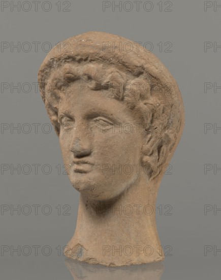 Female votive head, Female head in a wreath, slightly tilted to the right shoulder. Under the wreath are visible regularly arranged locks of hair, framing a low, triangular forehead and temples. Ears exposed. Deep-set eyes, almost flat in elaborately worked eyelids, small nose, small mouth with fleshy lips, short, strongly defined chin, full cheeks. 3rd century BC, Etruria. Votive. Height 26 cm, width 16.5 cm, depth 13.5 cm, weight 2.1 kg., A sculpted head made of a brownish clay-like material depicting a human face with short curly hair and a rounded cap or headband, showing worn surface texture and small chips, set against a uniform gray background; the features include a straight nose, closed lips, defined eyes with carved lids, and a long neck, with visible roughness and lighter and darker brown variations across the piece.