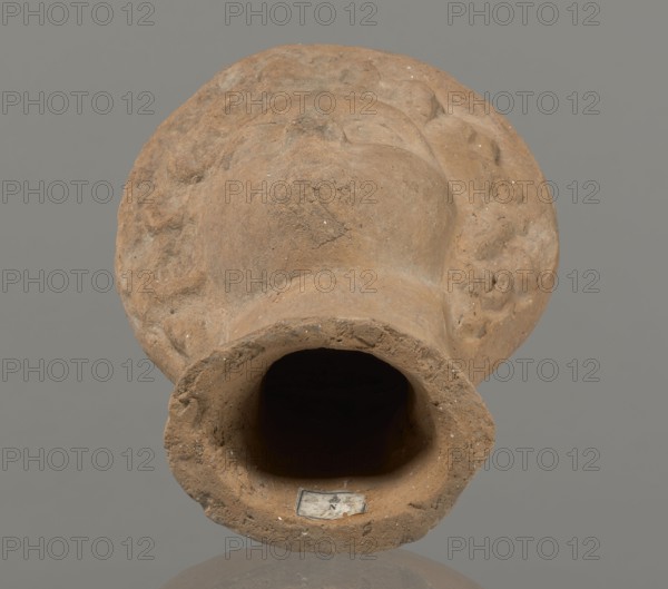 Female votive head, Female head in a wreath, slightly tilted to the right shoulder. Under the wreath are visible regularly arranged locks of hair, framing a low, triangular forehead and temples. Ears are exposed. Deep-set eyes, almost flat in the elaborately worked eyelids, small nose, small mouth with fleshy lips, short, strongly defined chin, full cheeks. 3rd century BC, Etruria; votive. Height 26 cm, width 16.5 cm, depth 13.5 cm, weight 2.1 kg., A weathered terracotta object resembling a hollow vessel or fragment with a rounded, flared opening and an irregular rim, showing a rough, granular surface texture and worn edges; the piece has molded, raised areas and shallow depressions around its exterior suggesting sculpted forms, and inside the opening there is a dark hollow. The visible colors are shades of brown and tan on the object, a dark interior, and a flat gray background.
