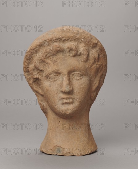 Female votive head, Female head in a wreath, slightly tilted to the right shoulder. Under the wreath are visible regularly arranged locks of hair, framing a low, triangular forehead and temples. Ears are exposed. Deep-set eyes, almost flat in the elaborately worked eyelids, small nose, small mouth with fleshy lips, short, strongly defined chin, full cheeks. 3rd century BC, Etruria. Votive. Height 26 cm, width 16.5 cm, depth 13.5 cm, weight 2.1 kg., A single sculpted head made of a rough, warm brown material sits against a smooth neutral gray background, showing a rounded hairstyle with textured curls, a defined forehead, closed or gently downcast eyes, a straight nose, and a subtle mouth, all with a weathered, pitted surface and small pale flecks; the neck is cylindrical and slightly flared at the base, and the overall tones include warm brown, light tan, small pale spots, and the surrounding gray.