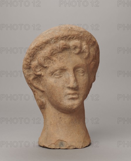 Female votive head, Female head in a wreath, slightly tilted to the right shoulder. Under the wreath are visible regularly arranged locks of hair, framing a low, triangular forehead and temples. Ears are exposed. Deep-set eyes, almost flat in the elaborately worked eyelids, small nose, small mouth with fleshy lips, short, strongly defined chin, full cheeks. 3rd century BC, Etruria, votive. Height 26 cm, width 16.5 cm, depth 13.5 cm, weight 2.1 kg., A weathered terracotta sculpted head displayed against a plain light gray background, showing a stylized face with closed or faintly defined eyes, a straight nose, and a slightly parted mouth; curly hair frames the forehead and temples and a rounded caplike form rests atop the head; the neck is elongated and cylindrical, ending in an uneven base that sits on the surface; the surface texture is rough with small pits and abrasion marks, and the visible colors include shades of warm brown, reddish-brown, tan, gray, and small specks of off-white...