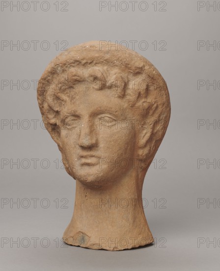 Female votive head, Female head in a wreath, slightly tilted to the right shoulder. Under the wreath are visible regularly arranged locks of hair, framing a low, triangular forehead and temples. Ears are exposed. Deep-set eyes, almost flat in the elaborately worked eyelids, small nose, small mouth with fleshy lips, short, strongly defined chin, full cheeks. 3rd century BC, Etruria; votive. Height 26 cm, width 16.5 cm, depth 13.5 cm, weight 2.1 kg., A sculpted head made of a brownish clay-like material sits against a smooth gray background, showing a rounded hairline with textured curls, a straight nose, closed or neutral eyes, and a slightly downturned mouth; the neck tapers to a flared base with some rough edges and small surface spots, and the visible color palette includes shades of brown, tan, and gray.