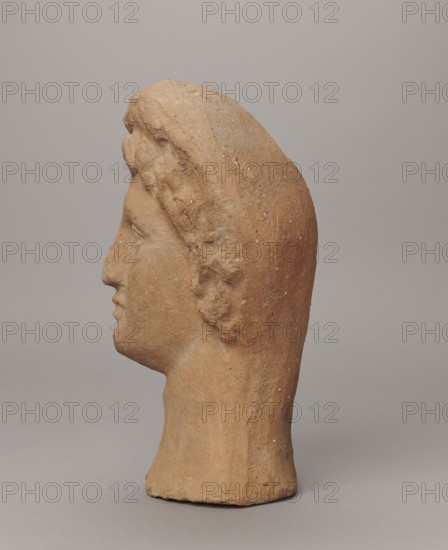 Female votive head, Female head in a wreath, slightly tilted to the right shoulder. Under the wreath are visible regularly arranged locks of hair, framing a low, triangular forehead and temples. Ears are exposed. Deep-set eyes, almost flat in the elaborately worked eyelids, small nose, small mouth with fleshy lips, short, strongly defined chin, full cheeks. 3rd century BC, Etruria; votive. Height 26 cm, width 16.5 cm, depth 13.5 cm, weight 2.1 kg., A weathered terracotta sculpture of a human head shown in profile with a pronounced nose, lips, and chin, textured hair forming a raised band along the top and back of the head, and a long neck extending to an uneven base; the surface shows rough, granular texture with small pits and imperfections. The visible colors are earthy brown and tan on the sculpture, against a smooth neutral gray background.