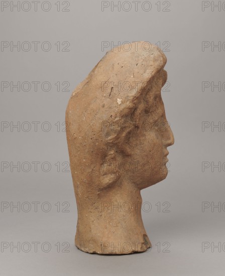 Female votive head. Female head in a wreath, slightly tilted to the right shoulder. Under the wreath are visible regularly arranged locks of hair, framing a low, triangular forehead and temples. Ears are exposed. Deep-set eyes, almost flat in the elaborately worked eyelids, small nose, small mouth with fleshy lips, short, strongly defined chin, full cheeks. 3rd century BC, Etruria. Votive. Height 26 cm, width 16.5 cm, depth 13.5 cm, weight 2.1 kg., A profile view of a terracotta-colored sculpted head with a textured surface and worn edges, showing the contour of a forehead, nose, lips, chin, and a stylized ear, set against a smooth light gray background with tones of brown, beige, and muted gray.