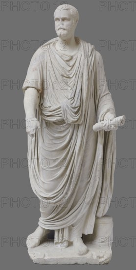 Roman statue, unknown Roman workshop. The figure of a standing man dressed in a tunic and toga, depicted in contrapposto. The right arm is exposed from the elbow, the left is almost completely hidden under the toga. Head from another statue. The face is small, with short stubble laid flat but carefully modeled. The mouth is compact, the corners turned slightly downward. The eyes are large, shadowed by drawn-down eyebrows, with a marked iris and pupil. Rectangular base, unprofiled. 1st–3rd century torso ca. mid-1st century AD; head 3rd quarter of 3rd century AD, Roman Empire 27 BC–476. Sculpture, weight 870 kg., A full-length white marble statue of a bearded man standing on a rectangular base, wearing layered draped garments with detailed folds that cover his body from shoulders to feet, one arm extended downward with the hand holding part of the drapery and the other arm bent forward holding a rolled object; the figure has short textured hair and a trimmed beard, a visible neckline ben...