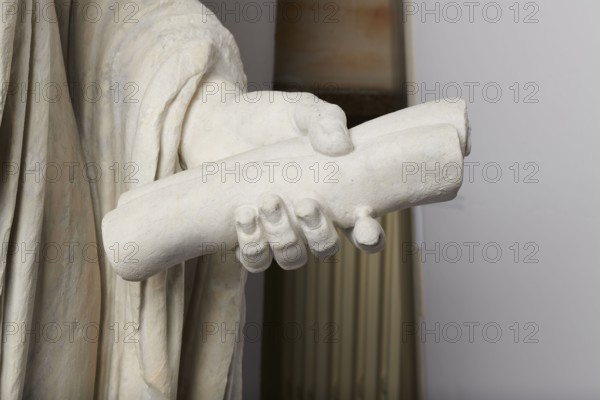 Roman statue, unknown Roman workshop. The figure of a standing man dressed in a tunic and toga, depicted in contrapposto. The right arm is exposed from the elbow, the left is almost completely hidden under the toga. Head from another statue. The face is small, with short stubble laid flat but carefully modeled. The mouth is compact, the corners turned slightly downward. The eyes are large, shadowed by drawn-down eyebrows, with a marked iris and pupil. Rectangular base, unprofiled. 1st–3rd century torso ca. mid-1st century AD; head 3rd quarter of 3rd century AD. Roman Empire 27 BC–476. Sculpture, weight 870 kg., A close-up of a pale off-white sculpted hand holding a cylindrical object, with draped fabric folds visible near the wrist; the sculpture shows textured surface details and rounded fingertips, set against a background featuring muted beige and gray vertical elements and a light gray wall, with colors including off-white, cream, beige, gray, and hints of greenish-beige on the dra...