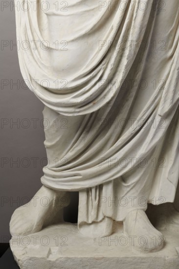 Roman statue, unknown Roman workshop. The figure of a standing man dressed in a tunic and toga, depicted in contrapposto. The right arm is exposed from the elbow, the left is almost completely hidden under the toga. Head from another statue. The face is small, with short stubble laid flat but carefully modeled. The mouth is compact, the corners turned slightly downward. The eyes are large, shadowed by drawn-down eyebrows, with a marked iris and pupil. Rectangular base, unprofiled. 1st–3rd century torso ca. mid-1st century AD; head 3rd quarter of 3rd century AD, Roman Empire 27 BC–476. Sculpture. Weight 870 kg., A close-up view of the lower portion of a carved stone statue showing draped fabric folds cascading over a base and covering legs and feet, with visible sculpted contours and creases; the material appears pale off-white with subtle gray and beige tones, and the background is a muted gray.