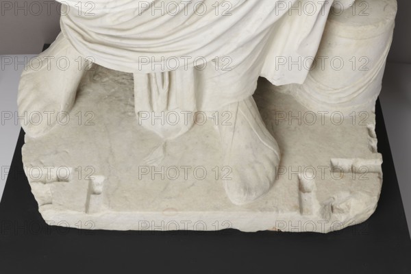 Roman statue, unknown Roman workshop. The figure of a standing man dressed in a tunic and toga, depicted in contrapposto. The right arm is exposed from the elbow, the left is almost completely hidden under the toga. Head from another statue. The face is small, with short stubble laid flat but carefully modeled. The mouth is compact, the corners turned slightly downward. The eyes are large, shadowed by drawn-down eyebrows, with a marked iris and pupil. Rectangular base, unprofiled. 1st–3rd century; torso ca. mid-1st century AD; head 3rd quarter of 3rd century AD. Roman Empire 27 BC–476. Sculpture. Weight 870 kg., A carved stone base with a draped cloth and a portion of a figure's foot and sandal, all in off-white and pale cream tones, resting on a black surface with small areas of light beige discoloration and subtle gray veining.