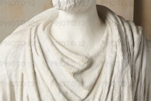 Roman statue, unknown Roman workshop. The figure of a standing man dressed in a tunic and toga, depicted in contrapposto. The right arm is exposed from the elbow, the left is almost completely hidden under the toga. Head from another statue. The face is small, with short stubble laid flat but carefully modeled. The mouth is compact, the corners turned slightly downward. The eyes are large, shadowed by drawn-down eyebrows, with a marked iris and pupil. Rectangular base, unprofiled. 1st–3rd century torso ca. mid-1st century AD; head 3rd quarter of 3rd century AD. Roman Empire 27 BC–476. Sculpture. Weight 870 kg., The image shows a close-up of a sculpture's upper torso and neck draped in fabric-like folds. The surface is predominantly white with subtle off-white and beige tones, and the texture varies from smooth areas on the neck to rougher, carved details on the draped sections. A trimmed beard is visible at the base of the chin, and the drapery forms layered, flowing folds across the c...