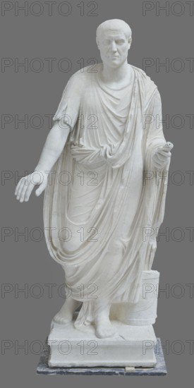 Roman statue, unknown Roman workshop. Figure of a striding man dressed in a tunic and toga. The folds of the toga flow widely, a slit at the left leg. The head is a large addition, with closely cropped hair and beard carved with short cuts of a pointed chisel. The face, with prominent jaws, tight mouth, and drawn eyebrows, is characterized by a stern expression. The pupils are marked. Profiled base, rectangular at the front, behind the back of the figure with rounded side edges. 2nd–3rd century torso 2nd century AD, head late 3rd century AD; earlier dating 1st–2nd century AD. Roman Empire 27 BC–476. Sculpture. Weight 928 kg., A white marble statue of a robed figure stands on a rectangular base against a gray background, showing draped fabric with flowing folds and layered textures over the shoulders, chest, and legs; the figure's right arm is extended outward with an open hand while the left arm is slightly bent holding a small cylindrical object, and the feet are bare with one foot sl...
