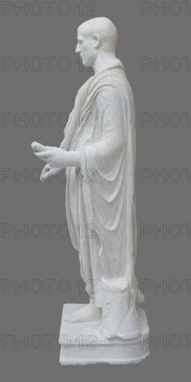 Roman statue, unknown Roman workshop. Figure of a striding man dressed in a tunic and toga. The folds of the toga flow widely, with a slit at the left leg. The head is a large addition, with closely cropped hair and beard carved with short cuts of a pointed chisel. The face, with prominent jaws, tight mouth, and drawn eyebrows, has a stern expression. The pupils are marked. Profiled base, rectangular at the front, behind the back of the figure with rounded side edges. Date: 2nd–3rd century torso 2nd century AD, head late 3rd century AD; earlier dating: 1st–2nd century AD. Roman Empire 27 BC–476. Sculpture. Weight 928 kg., A white marble statue of a robed male figure shown in profile against a solid gray background, with the figure standing on a rectangular base and wearing long, draped garments that form several folds and layers; the figure’s head is turned slightly forward, the hair is short and textured, the face shows a calm expression, and both arms are extended forward with hands ...