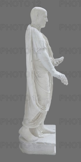 Roman statue, unknown Roman workshop. Figure of a striding man dressed in a tunic and toga. The folds of the toga flow widely, a slit at the left leg. The head is a large addition, with closely cropped hair on the head and beard, carved with short cuts of a pointed chisel. The face, with prominent jaws, tight mouth, and drawn eyebrows, is characterized by a stern expression. The pupils are marked. Profiled base, rectangular at the front, behind the back of the figure with rounded side edges. 2nd–3rd century torso 2nd century AD, head late 3rd century AD; earlier dating 1st–2nd century AD. Roman Empire 27 BC–476. Sculpture. Weight 928 kg., A white marble statue of a robed standing male figure shown in profile against a gray background, with carved drapery cascading from the shoulders down to the feet, a cloak hanging over one shoulder, an outstretched arm with a relaxed hand, the other hand slightly cupped near the torso holding a small rounded object, a short cropped hairstyle and defi...