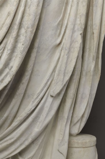 Roman statue, unknown Roman workshop. Figure of a striding man dressed in a tunic and toga. The folds of the toga flow widely, with a slit at the left leg. The head is a large addition, with closely cropped hair and beard carved with short cuts of a pointed chisel. The face has prominent jaws, a tight mouth, drawn eyebrows, and a stern expression; the pupils are marked. Profiled base, rectangular at the front, with rounded side edges behind the back of the figure. Torso 2nd century AD; head late 3rd century AD overall dated 2nd–3rd century; previously dated 1st–2nd century AD. Roman Empire 27 BC–476. Sculpture. Weight 928 kg., The image shows a close-up of draped fabric folds on a sculpture with a smooth, slightly textured surface in shades of off-white, cream, pale beige, and light gray, with subtle areas of warmer yellowish and faint brownish discoloration; the folds create overlapping curved lines and soft shadows, and a rounded cylindrical pedestal in similar off-white and beige to...