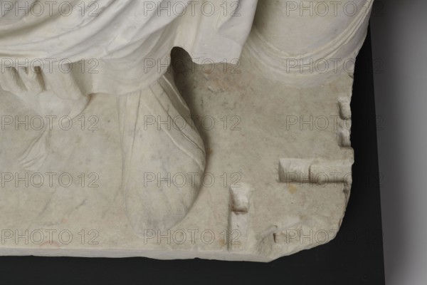 Figure of a striding man dressed in a tunic and toga. The folds of the toga flow widely, with a slit at the left leg. The head is a large addition, with closely cropped hair and beard carved with short cuts of a pointed chisel. The face has prominent jaws, a tight mouth, drawn eyebrows, and a stern expression; the pupils are marked. Profiled base, rectangular at the front and rounded at the sides behind the back of the figure. Roman statue, unknown Roman workshop. 2nd–3rd century torso 2nd century AD, head late 3rd century AD; earlier dating: 1st–2nd century AD. Roman Empire 27 BC–476. Sculpture. Weight 928 kg., The image shows a close-up of a carved pale stone surface with draped fabric and a rounded foot forming part of a statue, resting on a flat base. The stone appears mostly off-white with subtle gray veining and faint beige and tan discolorations. The drapery has carved folds and a few smooth curved lines, and the foot shows carved seams suggesting footwear. The base contains sev...