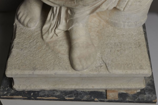Roman statue, unknown Roman workshop. Figure of a striding man dressed in a tunic and toga. The folds of the toga flow widely, a slit at the left leg. The head is a large addition, with closely cropped hair and beard, carved with short cuts of a pointed chisel. The face, with prominent jaws, tight mouth, and drawn eyebrows, is characterized by a stern expression. The pupils are marked. Profiled base, rectangular at the front, behind the back of the figure with rounded side edges. 2nd–3rd century torso 2nd century AD, head late 3rd century AD; earlier dating 1st–2nd century AD. Roman Empire 27 BC–476. Sculpture. Weight 928 kg., The image shows the lower portion of a sculpture resting on a rectangular stone base, including a carved foot and draped fabric. The stone surfaces display a textured, worn appearance with subtle variations of off-white, beige, and pale gray, and faint patches of light pinkish tones. The base sits on a dark, chipped platform with areas of black, brown, and expose...