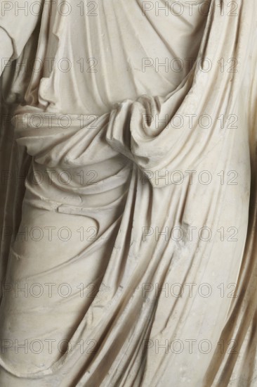 Roman statue, unknown Roman workshop. Figure of a striding man dressed in a tunic and toga. The folds of the toga flow widely, a slit at the left leg. The head is a large addition, with closely cropped hair on the head and beard, carved with short cuts of a pointed chisel. The face, with prominent jaws, tight mouth, and drawn eyebrows, is characterized by a stern expression. The pupils are marked. Profiled base, rectangular at the front, behind the back of the figure with rounded side edges. 2nd–3rd century torso – 2nd century AD; head – late 3rd century AD; earlier dating: 1st–2nd century AD. Roman Empire 27 BC–476. Sculpture. Weight 928 kg., A close-up view of draped fabric folds on a pale stone statue showing a section of torso and hip with flowing, gathered cloth; the surface is primarily off-white and cream with subtle beige and light gray shadows accentuating the carved creases and rounded shapes, and a few small areas of slightly darker discoloration and wear on the stone.