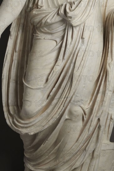Roman statue, unknown Roman workshop. Figure of a striding man dressed in a tunic and toga. The folds of the toga flow widely, with a slit at the left leg. The head is a later addition, with closely cropped hair and beard carved with short cuts of a pointed chisel. The face, with prominent jaws, tight mouth, and drawn eyebrows, has a stern expression; the pupils are marked. Profiled base, rectangular at the front, rounded side edges behind the figure. 2nd–3rd century torso 2nd century AD, head late 3rd century AD; earlier dating: 1st–2nd century AD. Roman Empire 27 BC–476. Sculpture. Weight 928 kg., A close-up of a carved stone draped figure showing detailed folds and flowing fabric in shades of off-white, cream, pale beige, and light gray, with subtle darker gray shadows and small areas of faint warm discoloration, revealing the sculpted contours of limbs beneath the cloth and the textured surface of the stone.