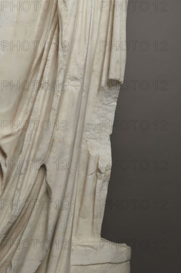 Roman statue, unknown Roman workshop. Figure of a striding man dressed in a tunic and toga. The folds of the toga flow widely, with a slit at the left leg. The head is a large addition, with closely cropped hair and beard carved with short cuts of a pointed chisel. The face, with prominent jaws, tight mouth, and drawn eyebrows, is characterized by a stern expression. The pupils are marked. Profiled base, rectangular at the front, with rounded side edges behind the back of the figure. 2nd–3rd century torso 2nd century AD; head late 3rd century AD; earlier dating 1st–2nd century AD. Roman Empire 27 BC–476. Sculpture. Weight 928 kg., The image shows a close-up of a carved stone surface with draped folds resembling fabric, displaying a smooth pale off-white to cream color with subtle warm beige and light brown discolorations and some darker grayish smudges; the carved folds create deep shadows and rounded ridges, and the right edge transitions to a rougher, broken texture with jagged protr...