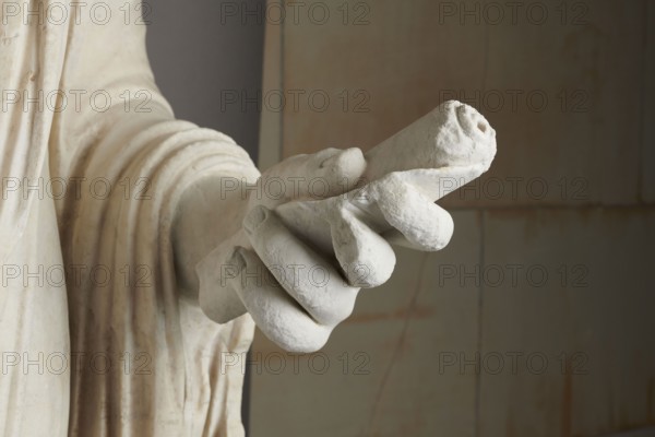 Roman statue, unknown Roman workshop. Figure of a striding man dressed in a tunic and toga. The folds of the toga flow widely, a slit at the left leg. The head is a large addition, with closely cropped hair on the head and beard, carved with short cuts of a pointed chisel. The face, with prominent jaws, tight mouth, and drawn eyebrows, is characterized by a stern expression. The pupils are marked. Profiled base, rectangular at the front, behind the back of the figure with rounded side edges. 2nd–3rd century torso – 2nd century AD, head – late 3rd century AD; earlier dating: 1st–2nd century AD. Roman Empire 27 BC–476. Sculpture. Weight 928 kg., A close-up of a carved stone hand holding a rolled cylinder, the hand and cylinder are off-white with subtle beige and light gray weathering, the hand shows rounded fingers and textured surfaces, the wrist and draped sleeve beside it display carved folds and a creamy off-white color with faint brownish stains, the background is a muted mix of gra...