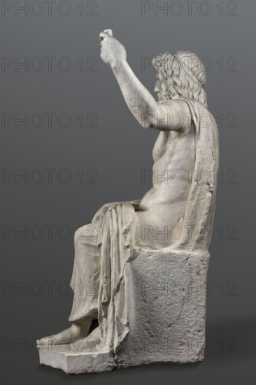 Statue of Jupiter of the Capitoline type, unknown Roman workshop. Jupiter Zeus seated on a throne set on a rectangular base. Roman sculpture, Roman copy after a Hellenistic variant of Phidias's statue of Zeus the Olympian. Zeus sits stiffly with his torso slightly back; his raised left hand holds a scepter and his right hand rests on his right knee. Right foot slightly forward. Legs covered with a himation that also covers the back and left shoulder. Muscular torso, strongly developed shoulders, with naturalistic modeling. Head disproportionately large; face with slightly open mouth. Hair parted from the center anastole. Thick beard arranged in strands on the sides and in curls on the chin. Late 2nd–3rd century AD. Roman Empire 27 BC–476 AD. Sculpture. Weight 930 kg., A white marble statue of a seated male figure viewed in profile against a gray background, showing a few flowing locks of hair and a beard, an arm raised with a hand holding an unseen object, a draped garment over the sho...