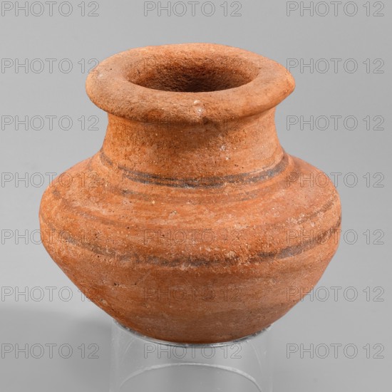 Vessel, A small wheel-made clay vessel with an almost hemispherical belly that transitions halfway up into clearly defined shoulders and a rounded bottom. Short neck tapering toward the top and an upturned rim. Interior surface smoothed; exterior smoothed and covered with an orange engobe. Decoration: four horizontal bands — two on the lower part of the neck and two on the body. Vessel complete, slightly eroded, especially on the neck. Likely early 18th Dynasty, ca. 1550–1295 BC New Kingdom, 18th Dynasty. Type: vessel. Height 7 cm; width 7.5 cm; rim diameter 5.1 cm; weight 0.12 kg. Possible marl clay type MA4? per Vienna System classification., A rounded earthenware vessel with a wide, flared rim and a narrow neck expanding into a bulbous body, shown against a plain light gray background; the vessel is textured and predominantly terracotta orange with a few darker brownish-gray horizontal bands and some lighter speckles, and it rests on a clear cylindrical stand.
