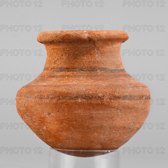 Vessel. A small wheel-made clay vessel with an almost hemispherical belly that transforms halfway up into clearly defined shoulders and a rounded bottom. Short neck tapering toward the top with an upturned rim. Interior smoothed; exterior smoothed and covered with an orange engobe. Decoration: four horizontal bands—two on the lower neck and two on the body. Vessel complete, slightly eroded, especially on the neck. Material: marl clay type MA4? according to Vienna System. Function: tableware. Painted pottery, Egyptian ceramics. Early 18th Dynasty?, ca. 1550–1295 BC New Kingdom, 18th Dynasty. Height 7 cm; width 7.5 cm; rim diameter 5.1 cm; weight 0.12 kg., A single terracotta vessel with a rounded body, a narrowed neck, and a flared rim sits on a clear cylindrical stand; the surface shows a coarse, textured finish with a few thin darker bands encircling the neck and upper body, and the visible colors include shades of orange, reddish-brown, darker brown in the bands, and neutral gray fro...