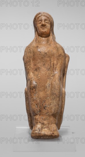 Figurine of a seated woman. Figurine of a woman sitting on a throne with her hands placed on her knees, made of reddish-yellowish clay. The frontally shown female figure has more clearly defined facial contours in relation to the body. She is dressed in a chiton and a himation thrown over the back of her head. The back of the figurine is unframed. East Greek Group type, 1st half of the 5th century BC previous dating: mid-5th century BC. Cyrene Libya. Terracotta figurine; height 9.5 cm, width 4 cm, depth 5.8 cm, weight 0.10 kg., A worn figurine carved from a light brown material, seated upright on a simple base with a few surface cracks and patches of darker brown discoloration, showing a rounded head with subtle facial features and a hood or close-fitting covering over the head, arms rested along the sides and hands suggested near the knees, the overall shape is smooth and slightly glossy in places with small areas of darker speckling and weathering, set against a neutral gray backgrou...