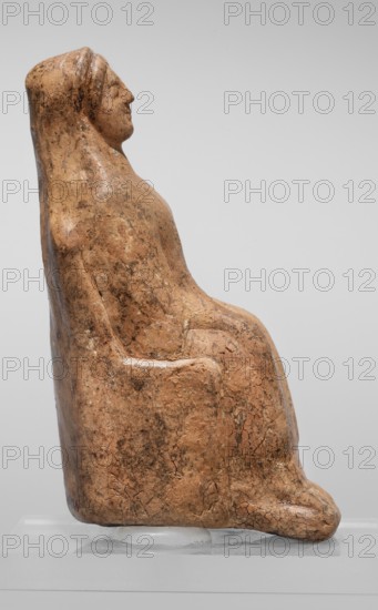 Figurine of a seated woman. Figurine of a woman sitting on a throne with her hands placed on her knees, made of reddish-yellowish clay. The frontally shown female figure has more clearly defined facial contours in relation to the body. She is dressed in a chiton and a himation thrown over the back of her head. The back of the figurine is unframed. The figurine is of the East Greek Group type, 1st half of the 5th century BC, Cyrene Libya. Figurine; height 9.5 cm, width 4 cm, depth 5.8 cm, weight 0.10 kg., A seated figurine carved from a brownish-beige material with a weathered, cracked surface and darker brown mottling, shown in profile; the figure has a rounded head with a subtle nose and chin, a smooth hood or draped shape extending down the back, a simplified torso and arms folded or resting at the sides, and legs tucked forward with a suggestion of feet, all resting on a clear display support against a pale gray background.