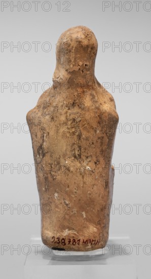 Figurine of a seated woman. Figurine of a woman sitting on a throne with her hands placed on her knees, made of reddish-yellowish clay. The frontally shown female figure has more clearly defined facial contours in relation to the body. She is dressed in a chiton and a himation thrown over the back of her head. The back of the figurine is unframed. The figurine is of the East Greek Group type, 1st half of the 5th century BC, Cyrene Libya. Figurine; height 9.5 cm, width 4 cm, depth 5.8 cm, weight 0.10 kg., A worn, upright figurine seen from the back with a rounded head and sloping shoulders, showing a rough, pitted surface with areas of discoloration and small surface deposits; the object is predominantly tan and brown with darker brown spotting and lighter beige patches, and it rests on a transparent stand against a pale gray background.